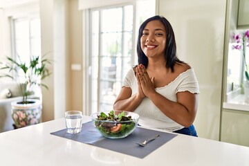 Obraz na płótnie Canvas Young hispanic woman eating healthy salad at home praying with hands together asking for forgiveness smiling confident.