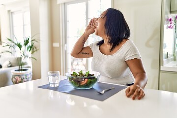 Young hispanic woman eating healthy salad at home shouting and screaming loud to side with hand on mouth. communication concept.