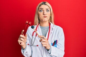 Young caucasian woman wearing doctor uniform holding tuning fork puffing cheeks with funny face....