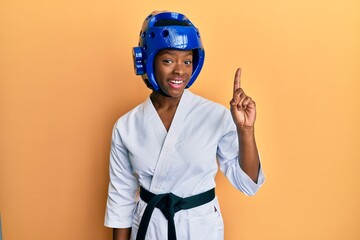 Young african american girl wearing taekwondo kimono and protection helmet smiling with an idea or question pointing finger up with happy face, number one