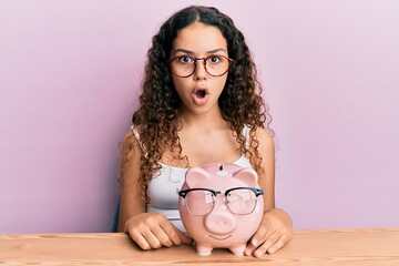 Obraz na płótnie Canvas Teenager hispanic girl holding piggy bank with glasses scared and amazed with open mouth for surprise, disbelief face
