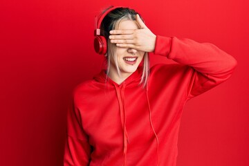 Young modern girl wearing gym clothes and using headphones smiling and laughing with hand on face covering eyes for surprise. blind concept.