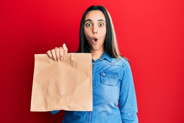 Fototapeta na wymiar Young hispanic woman holding take away paper bag scared and amazed with open mouth for surprise, disbelief face