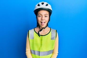 Beautiful brunette little girl wearing bike helmet and reflective vest sticking tongue out happy with funny expression. emotion concept.