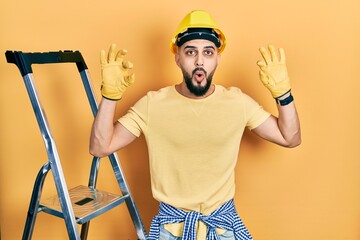 Handsome man with beard by construction stairs wearing hardhat looking surprised and shocked doing ok approval symbol with fingers. crazy expression