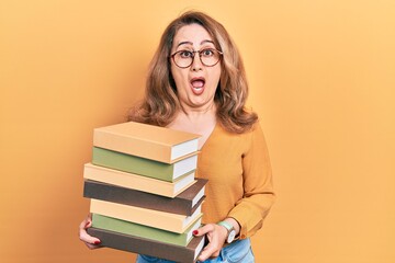 Middle age caucasian woman holding a pile of books afraid and shocked with surprise and amazed expression, fear and excited face.