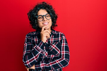 Fototapeta na wymiar Young hispanic woman with curly hair wearing casual clothes and glasses looking confident at the camera with smile with crossed arms and hand raised on chin. thinking positive.