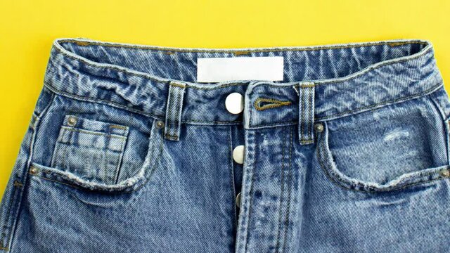 Moving picture of blue unisex jeans with metal buttons, zipper, pockets, white label flat lay on yellow background, copy space. Daily fashion clothes in minimalism style. Casual outfit. Denim jeans