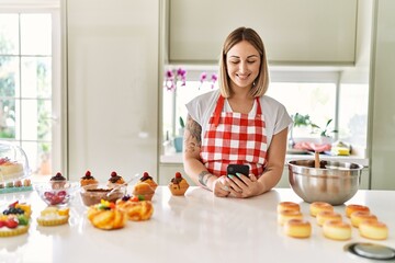 Young beautiful blonde woman wearing apron cooking pastries looking for recipe on smartphone looking positive and happy standing and smiling with a confident smile showing teeth