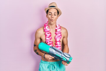 Young hispanic man wearing swimsuit and watergun making fish face with mouth and squinting eyes, crazy and comical.