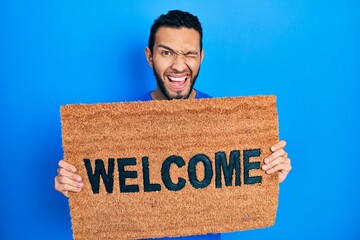 Hispanic man with beard holding welcome doormat winking looking at the camera with sexy expression,...