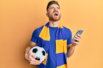 Young redhead man wearing hooligan scarf and using smartphone angry and mad screaming frustrated and furious, shouting with anger looking up.