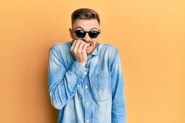 Young redhead man wearing stylish sunglasses looking stressed and nervous with hands on mouth biting nails. anxiety problem.