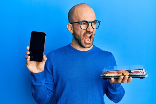 Young hispanic man holding take away food showing smartphone screen angry and mad screaming frustrated and furious, shouting with anger. rage and aggressive concept.