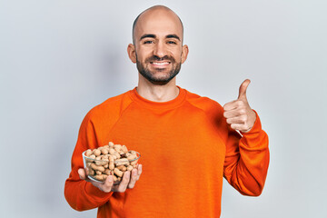 Young bald man holding peanuts smiling happy and positive, thumb up doing excellent and approval sign