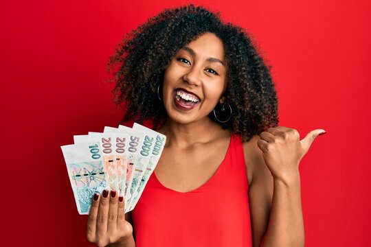 Beautiful african american woman with afro hair holding czech koruna banknotes pointing thumb up to the side smiling happy with open mouth