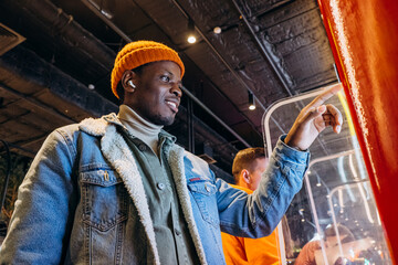 Smiling African-American man in warm denim jacket with wireless earphones uses self-service kiosk...