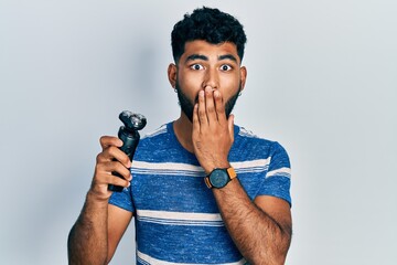 Arab man with beard holding electric razor machine covering mouth with hand, shocked and afraid for...