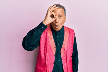 Middle age indian man wearing traditional indian clothes doing ok gesture shocked with surprised face, eye looking through fingers. unbelieving expression.