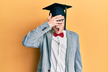 Young caucasian nerd man wearing glasses and graduation cap peeking in shock covering face and eyes with hand, looking through fingers afraid