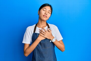 Young chinese woman wearing waiter apron smiling with hands on chest with closed eyes and grateful gesture on face. health concept.