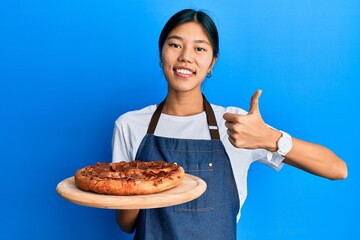 Young chinese woman wearing waiter apron holding italian pizza smiling happy and positive, thumb up...