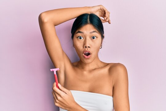 Young chinese woman wearing shower towel using razor afraid and shocked with surprise and amazed expression, fear and excited face.