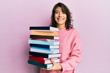 Young hispanic woman holding a pile of books smiling with a happy and cool smile on face. showing teeth.