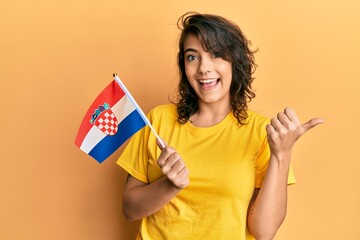 Young hispanic woman holding croatia flag pointing thumb up to the side smiling happy with open mouth