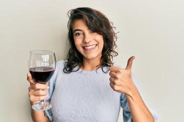 Young hispanic woman drinking a glass of red wine smiling happy and positive, thumb up doing excellent and approval sign