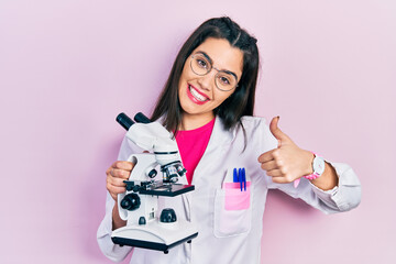 Young hispanic girl wearing scientist uniform holding microscope smiling happy and positive, thumb up doing excellent and approval sign