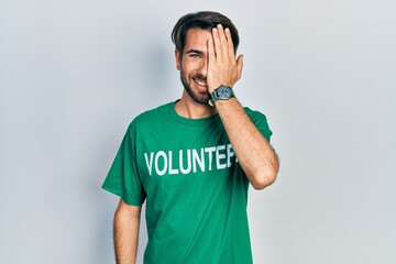 Young hispanic man wearing volunteer t shirt covering one eye with hand, confident smile on face...