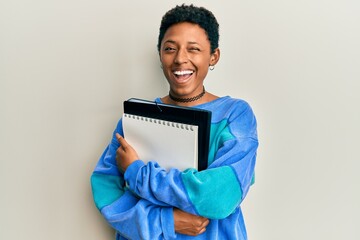 Young african american girl holding books winking looking at the camera with sexy expression,...