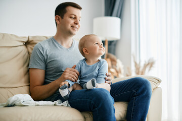 Cute baby boy and his father relaxing on the sofa at home. Smiling father enjoying with his baby boy who is sitting on his lap.