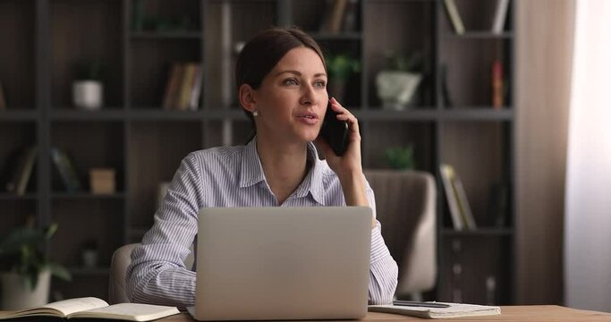 Young business woman sit at workplace desk look at laptop screen talking to assistant on cell phone discuss purchase, get consultation, buy online. Make order or negotiate to client distantly concept