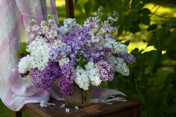 A bouquet of white, pink and purple flowers of lilac and gillyflower in a vase on a chair in the garden against the background of grass and bushes, sunlight, blur. Beautiful card. - Powered by Adobe