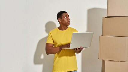Professional male mover in yellow t shirt looking aside, using laptop, standing near a stack of cardboard boxes while doing home relocation