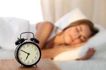 Beautiful young woman sleeping and smiling while lying in bed comfortably and blissfully on the background of alarm clock is going to ring. Sunbeam dawn on her face.