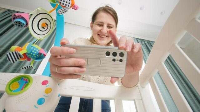 Happy smiling mother making video of her baby lying in cradle for social media. Concept of parenting and making images of children