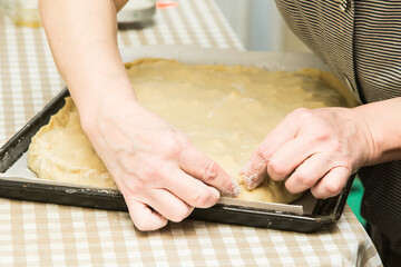 The hands of an adult elderly woman are preparing a dough pie. The fingers of the hands fasten the edges of the dough to each other.