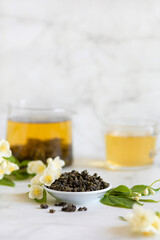 Chinese large leaf green tea with jasmine on white marble background, copy space, text. Dry leaves and brewed herbal drink in teapot and cup. vertical