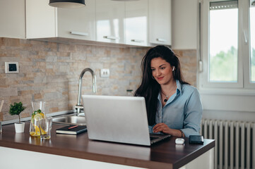 Woman using a laptop and airpods while sitting in the kitchen
