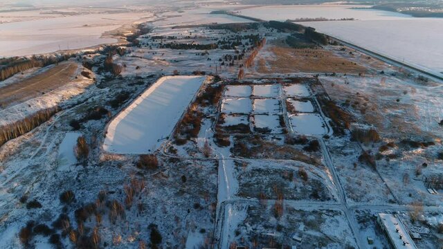 Purifying constructions of wastewater treatment plant on snowy land among fields against lake at sunset light aerial panorama