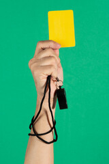 referee's hand holding a yellow card and whistle on green background.