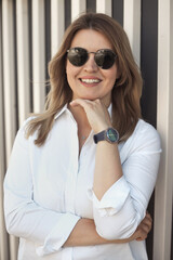 Lifestyle portrait of a smiling young confident woman 30-40 years old in a white shirt, wears sunglasses and a wrist watch