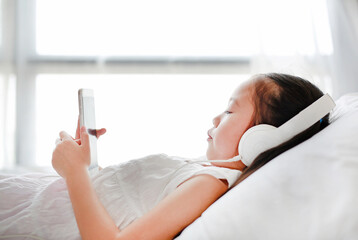 Portrait little Asian girl using headphones listen music by smartphone while lying on bed in bedroom at home.