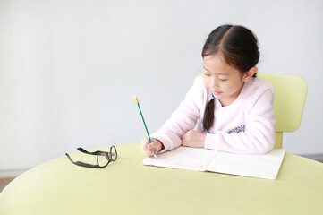 Portraits of little Asian girl writes in a book or notebook with pencil sitting on kid chair and table in classroom.