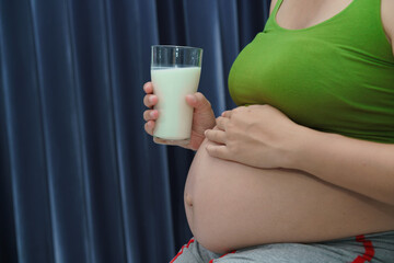 Close-up pregnant woman holding glass of milk sitting on sofa in the room. Nutrition and diet during pregnancy.