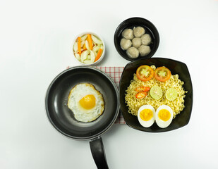 Indonesia Warm noodles served with eggs and fresh tomatoes with meatballs on a black bowl and pickled on a white bowl and eggs on a white background