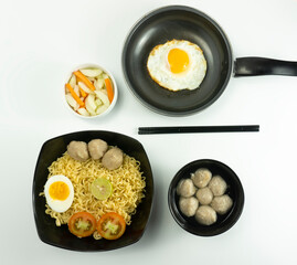 Indonesia Warm noodles served with eggs and fresh tomatoes with meatballs on a black bowl and pickled on a white bowl and eggs on a white background